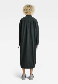 Robe noire à manches longues avec une coupe droite, dotée de fentes latérales pour faciliter les mouvements. Tissu lisse avec un col et une fermeture à boutons dans le dos.
