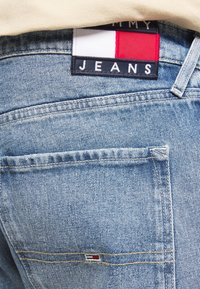 Blå denimjeans med en synlig logotypmärke i rött, vitt och marinblått. Två bakfickor med kontrasterande sömnadsdetaljer.