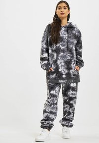 Felpa con cappuccio grigia e nera a tie-dye con tasca frontale, abbinata a pantaloni jogger coordinati. Il tessuto sembra morbido, con polsini elasticizzati e cappuccio.
