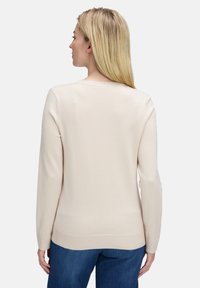 Hosszú szőke hajjal rendelkező nő, világos krémszínű, hosszú ujjú pulóvert és kék farmert visel, háttal áll egy világos, egyszínű háttér előtt.