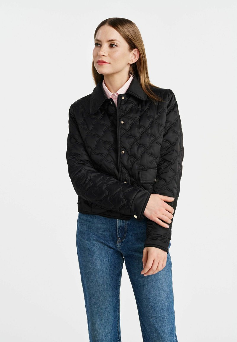 Veste matelassée noire avec col, boutons-pression et manches longues. Portée sur une chemise rose clair et associée à un jean bleu.