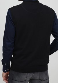 Czarna dzianinowa kamizelka z wysokim kołnierzem, prążkowanym dołem i gładkimi szwami, noszona na ciemnoniebieskiej koszuli z długim rękawem.