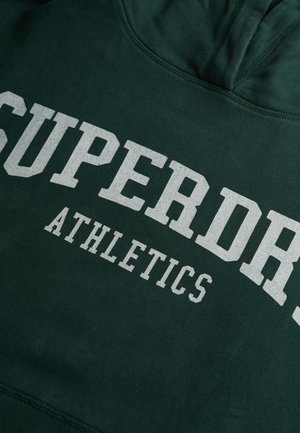 Ciemnozielona bluza z kapturem z białym, pogrubionym napisem "SUPERDRY ATHLETICS" na piersi, w czcionce w stylu uniwersyteckim.