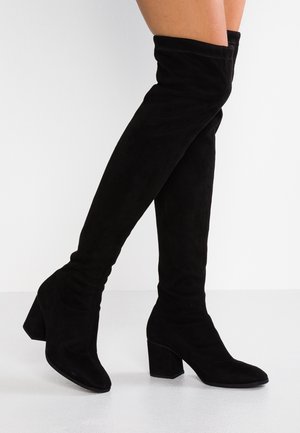 Czarne zamszowe kozaki za kolano na masywnym obcasie, noszone na skrzyżowanych nogach na białym tle.