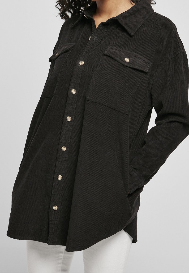 Chemise en velours côtelé noir avec un col, deux poches avant et une fermeture à boutons. Le tissu présente une texture côtelée et une coupe ample.