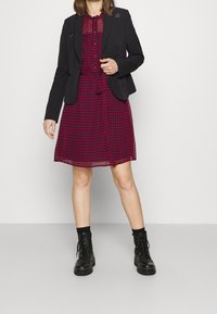 Černý blazer přes červené kostičkové šaty s vázaným pasem. Šaty mají přední zapínání na knoflíky a průsvitnou horní část. Černé šněrovací boty.