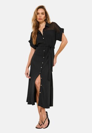 Femme portant une robe mi-longue noire à boutons sur le devant, avec des manches supérieures transparentes, une ceinture à la taille et des talons noirs à lanières, posant devant un fond blanc.