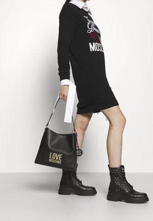 Zwarte gebreide jurk met witte kraag, voorzien van geborduurde tekst. Zwarte leren handtas met "LOVE MOSCHINO" in goud en sierlijke hardware. Zwarte veterlaarzen.