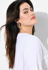 Haut blanc à manches courtes avec une coupe décontractée, présentant une texture lisse. Le sujet a les cheveux longs et ondulés, agrémentés de grandes boucles d'oreilles en créole.