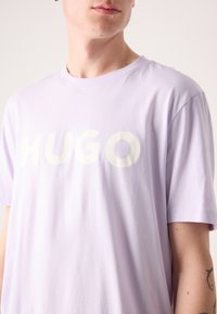 T-shirt en coton lavande clair avec un grand logo textuel "HUGO" blanc sur la poitrine. Col rond et manches courtes. Texture lisse.