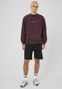 Jeune homme aux cheveux blonds courts portant un sweat-shirt violet avec le texte "Out all Day", un short noir, des chaussettes blanches et des baskets grises sur fond blanc.