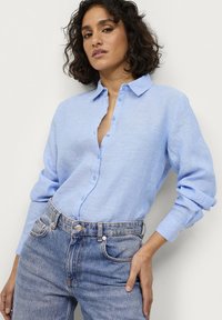 Camisa de lino azul claro con cuello abotonado, botones frontales y mangas largas. Combinada con pantalones vaqueros de mezclilla azul de tiro alto.