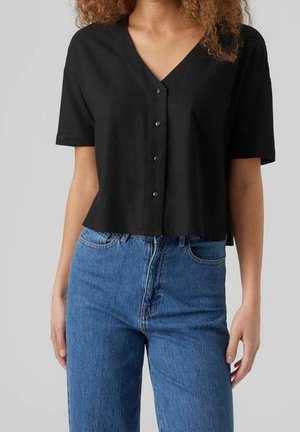 Woman wearing a black short-sleeve button-up V-neck top and high-waisted blue jeans against a plain background.