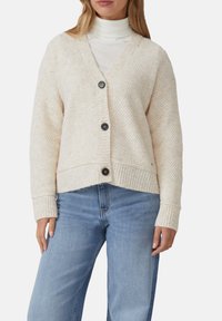 Frau in einem cremefarbenen, strukturierten, mit Knöpfen versehenen Cardigan über einem weißen Rollkragenpullover und hellblauen Jeans, die mit entspannten Armen dasteht.