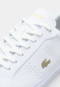 Baltos odinės sportbačių su auksiniu Lacoste logotipu, plokščiais baltais raišteliais, perforuotais detalėmis šone, lygiu paviršiumi, žemu profiliu.