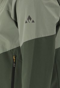 Veste verte avec une finition mate et texturée. Présente un logo sombre sur l'épaule et une poche zippée avec un accent jaune sur le côté.