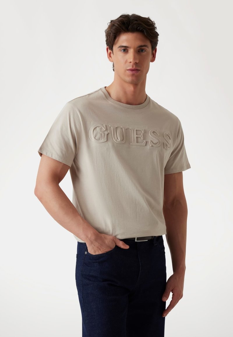 Camiseta de algodón beige con el logo "GUESS" en relieve en el frente, cuello redondo, mangas cortas, combinada con jeans azul oscuro.