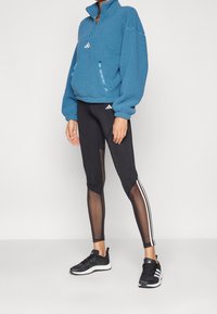 Blå fleecepullover med halvdragkedja, två sidofickor; svarta leggings med genomskinliga paneler och vita ränder; svarta sneakers.