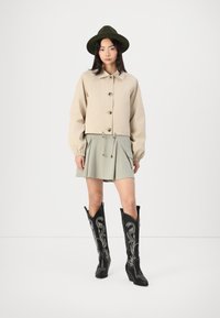 Beige kort jasje met knopen, groene geplooide rok en zwarte geborduurde cowboylaarzen, gestyled met een bijpassende groene hoed.
