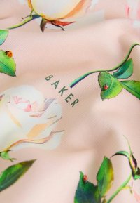 Kvetinová tkanina s jemne ružovým pozadím a bielymi a žltými ružami. Text "BAKER" je vytlačený na zeleno medzi zelenými listami.