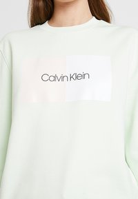 Světlé zelené tričko s velkým potištěným logem s nápisem „Calvin Klein“ černou barvou na obdélníkovém bílém a pastelově růžovém pozadí.