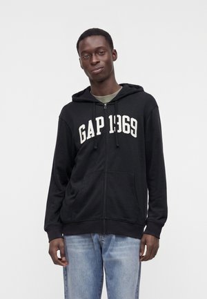 Joven con una sudadera con capucha negra con cremallera con el texto GAP 1969 y vaqueros azul claro, de pie contra un fondo liso y claro.