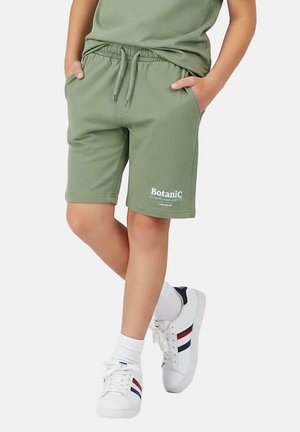 Groene katoenen korte broek met een elastische tailleband, zijzakken en de opdruk "Botanic" in het wit. Draagt witte sneakers.