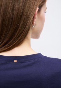 Γυναίκα με καστανά ίσια μαλλιά, που φορά ασημένιο σκουλαρίκι κρίκο και μπλε σκούρο πουκάμισο, εμφανιζόμενη από πίσω και πλάγια σε λευκό φόντο.