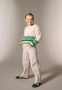 Jeune fille souriante avec les mains dans les poches, portant un pull en tricot crème et vert, un pantalon blanc et des baskets vertes, se tenant sur un fond neutre.
