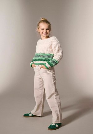 Young girl smiling with hands in pockets, wearing a cream and green knitted sweater, white pants, and green sneakers, standing on a neutral background.