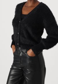 Czarny futrzany kardigan z dekoltem w serek, długimi rękawami i zapięciem na guziki z przodu. Połączony z gładkimi czarnymi skórzanymi spodniami. Miękka faktura, przytulny design.