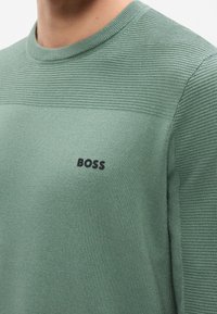 Pull vert texturé à manches longues avec nervures horizontales sur la poitrine et les épaules, arborant un logo noir "BOSS" sur le côté gauche de la poitrine.