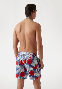 Gėlėtos maudymosi šortai su mėlynu fonu, kuriame raudoni hibiskų žiedai ir žali palmės lapai, elastinė juosmens juosta ir šoniniai kišeniai.