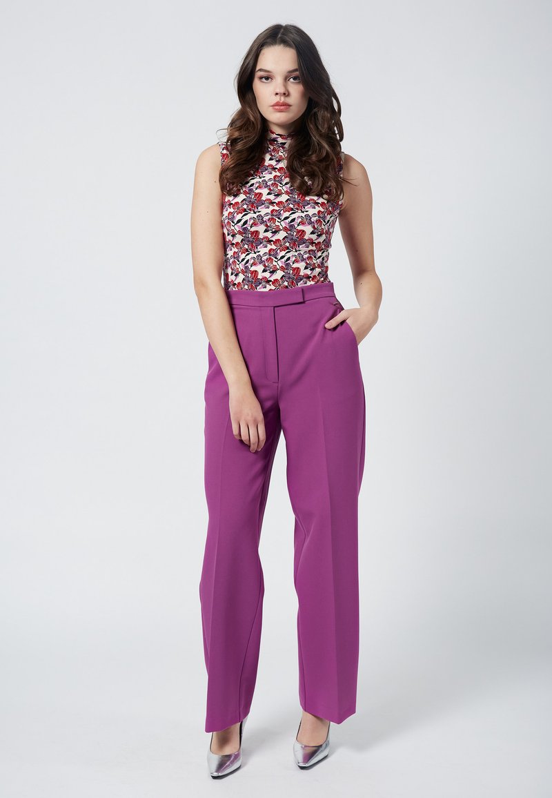 Blommig ärmlös topp och högmidjade lila byxor med skräddarsydd passform. Silverfärgade högklackade skor fulländar outfiten mot en enkel bakgrund.
