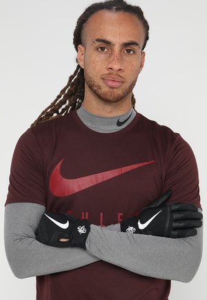 Svarte treningshanske med teksturert overflate, med en hvit logo. Brukt av en person i en burgunder Nike skjorte og grå langermet genser.