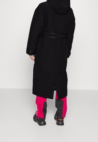 Svart lång kappa med huva, bälte i midjan och bakslits. Matchad med rosa och grå byxor samt svarta skor. Minimalistisk design.