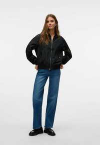 Veste bomber noire avec fermeture éclair à l'avant, poignets élastiques et poches latérales, associée à un jean droit bleu et des chaussures noires. Texture lisse et brillante.