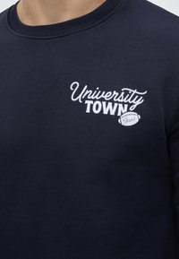 Μπλούζα σε ναυτικό μπλε με λευκό κείμενο "University Town" και μικρό γραφικό ποδοσφαίρου με "Gant" στο στήθος.