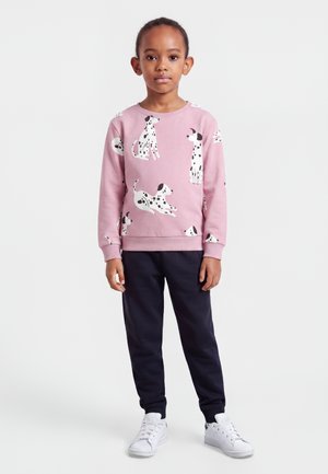 Enfant debout portant un sweat-shirt rose avec des imprimés de chiens dalmatiens, un pantalon noir et des baskets blanches sur un fond blanc uni.