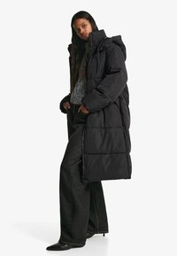 Svart vadderad jacka med avtagbar huva, dragkedja fram och quiltat mönster. Kombinerad med mörka byxor och en grå stickad tröja.