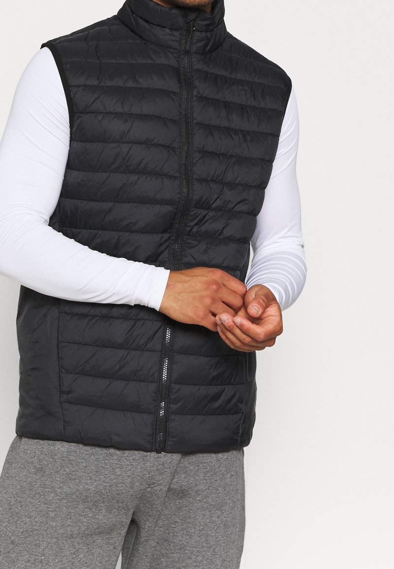 Homme portant un gilet matelassé noir sans manches par-dessus une chemise blanche à manches longues et un pantalon de survêtement gris, les mains jointes près de la taille.