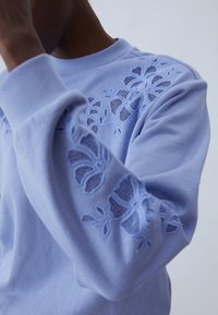 Osoba nosící světle fialovou mikinu s květinovou výšivkou s výřezy na rameni a rukávu, která zvedá ruku blízko obličeje.