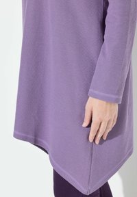 Fioletowa teksturowana tunika z długimi rękawami, z zaokrąglonym dołem i widocznymi detalami przeszyć. Materiał ma prążkowany wzór.