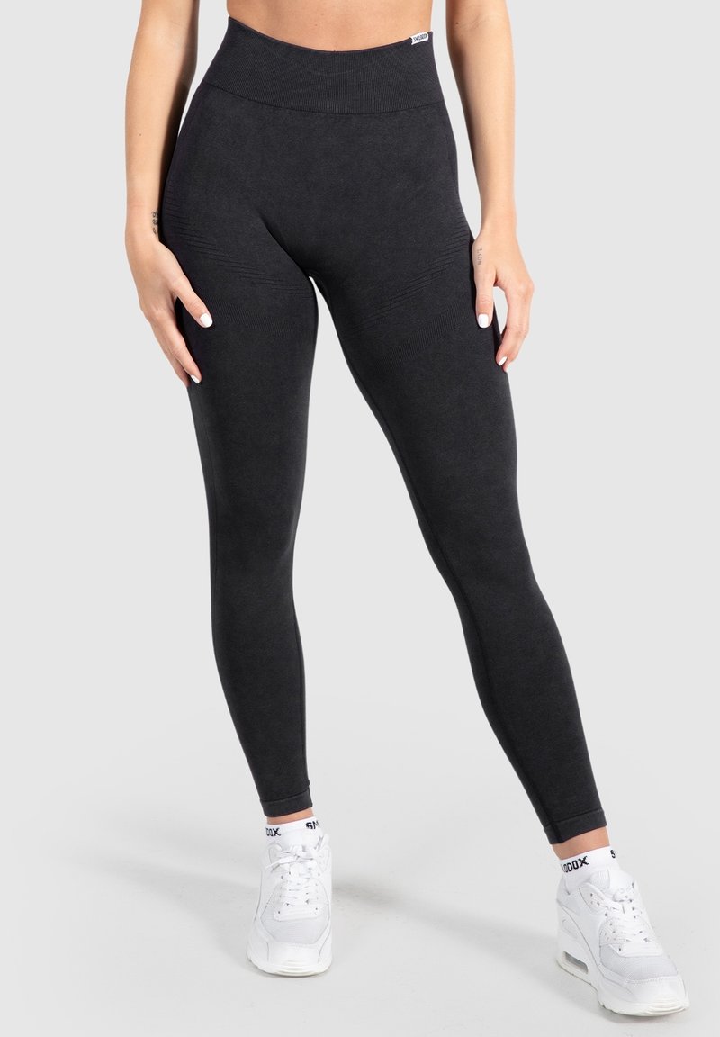 Femme portant des leggings noirs taille haute et des baskets blanches, debout les mains posées sur les cuisses contre un fond clair uni.
