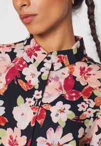 Femme aux cheveux tressés portant une chemise à fleurs boutonnée, avec des fleurs roses, rouges et blanches sur un fond noir.