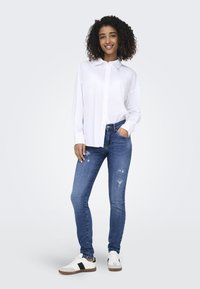 Camisa blanca de botones, jeans azules desgastados y zapatillas blancas con acentos negros. El modelo está de pie con una mano en el bolsillo, sonriendo.