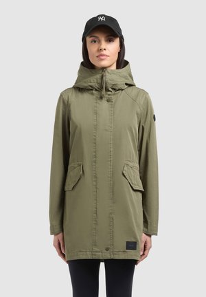 Nő, aki oliva zöld kapucnis dzsekit visel első zsebekkel és fekete baseball sapkával, világos, egyszerű háttér előtt áll.