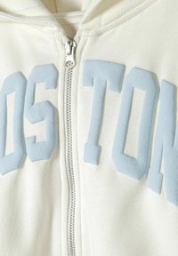 Cream zip-up hoodie featuring raised light blue "BOSTON" lettering, metal zipper, and soft textured fabric.