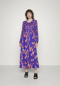 Vestido maxi de manga longa, floral, em roxo com padrões em laranja e azul. Tecido suave, decote redondo e silhueta fluída. Sapatos pretos de dedo aberto.