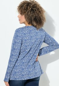 Donna con capelli ricci che indossa una camicia a maniche lunghe blu con motivi floreali bianchi, in piedi con una mano sui fianchi su uno sfondo bianco.